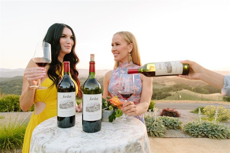 two women standing near high-top with two bottles of jordan cabernet sauvignon on table and glasses of cabernet in their hands