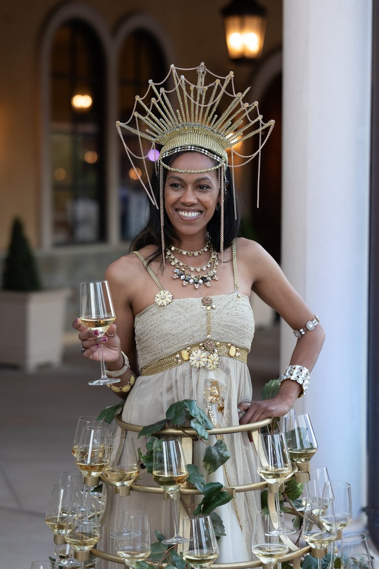 a wine server in a costume of a tiered metal skirt with glasses of wine in slots in the tiers and an elaborate crown headpiece