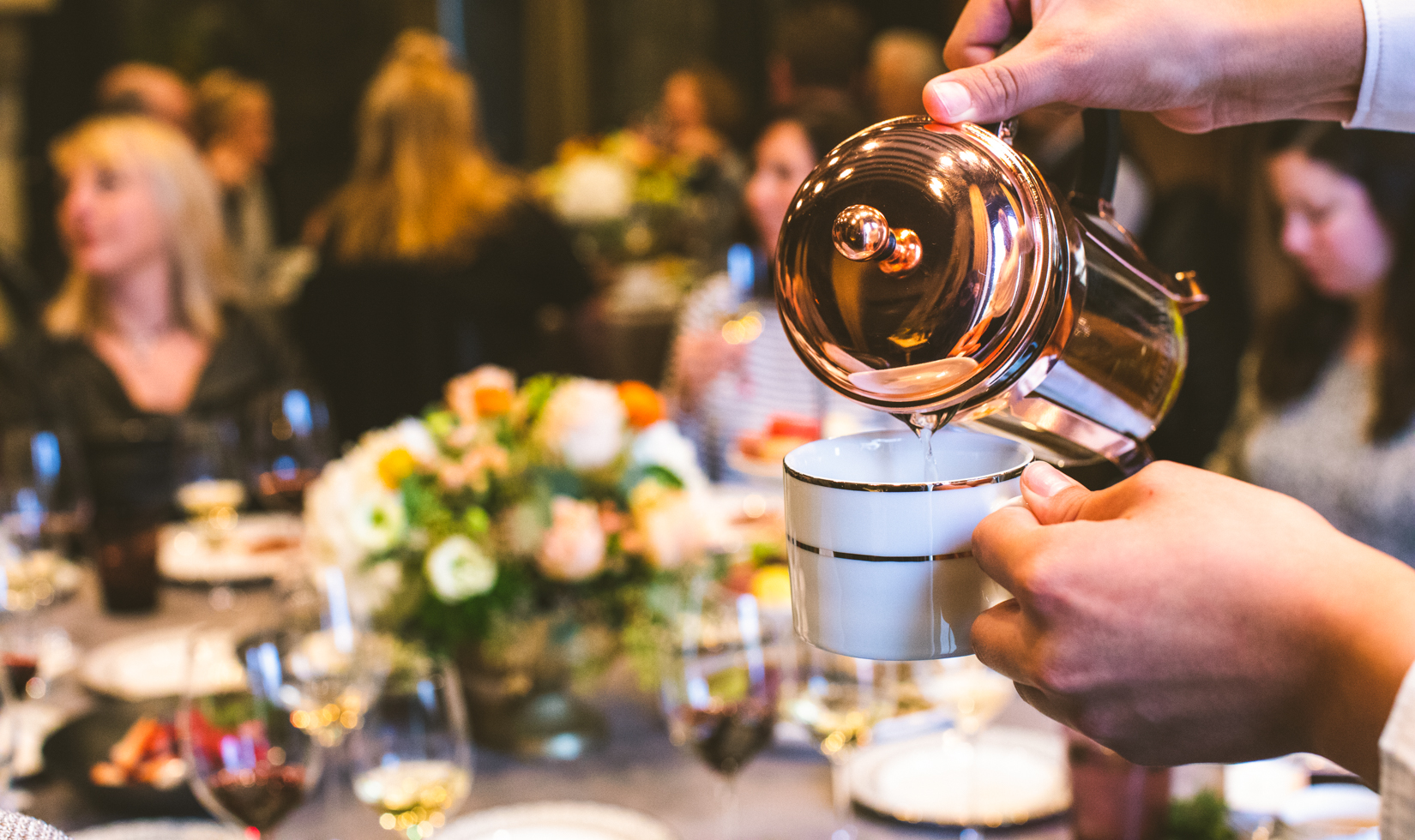 tea being poured into a small mug with group of seated people behind